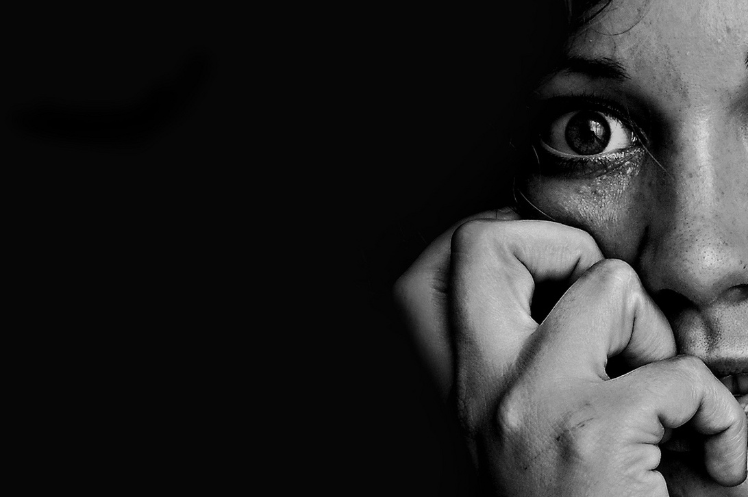 Ett svartvitt porträtt i hög kontrast som visar ett delvis synligt ansikte med ett öga som uttrycker rädsla. Händerna täcker munnen i en skyddande gest, vilket skapar en stark bild av ångest och emotionell stress.
