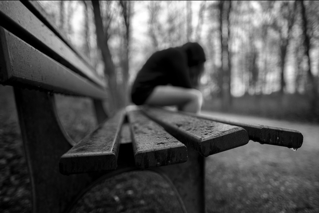 Ett stämningsfullt svartvitt fotografi som visar en bänk i ett parkliknande område med en suddig, hopkrupen gestalt på. Förgrunden fokuserar på bänkens träribbor medan bakgrunden visar kala träd. Kompositionen och det selektiva fokuset skapar en stark känsla av isolering och melankoli.